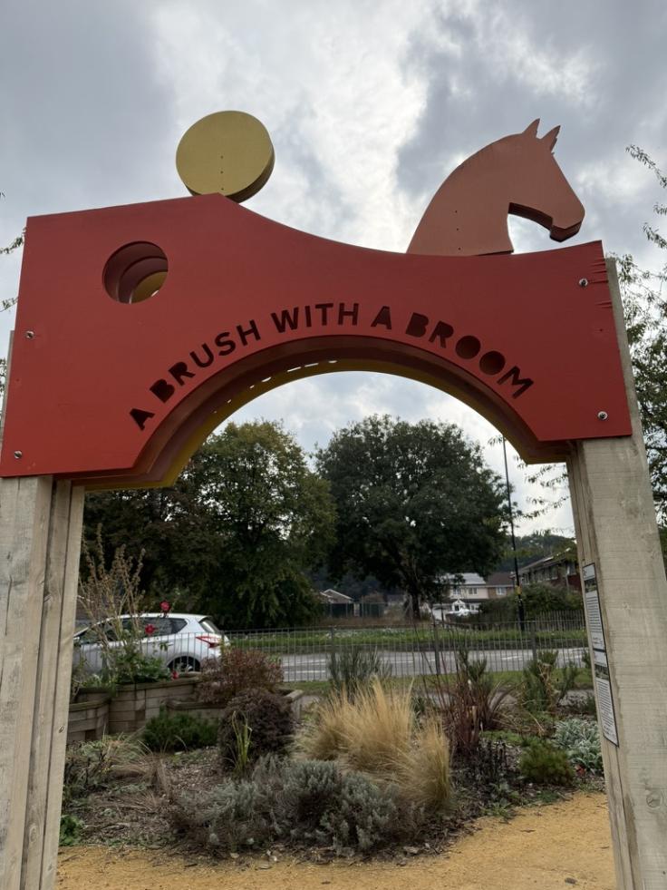 A metal archway with the words 'A brush with a broom' cut out across the span, and a horse's head silhouette on top.