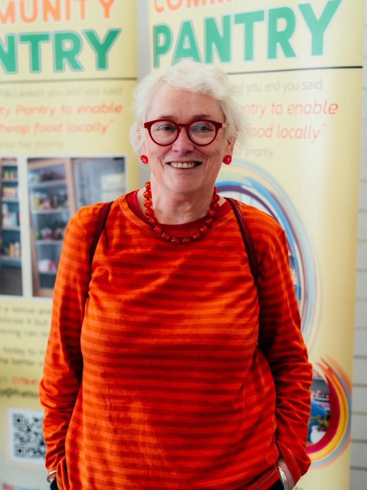 An older woman with white hair, red glasses, red earrings, a red necklace, a red and orange stripey top and her hands in her pockets smiles at the camera. She stands in front of some yellow pop-up signs advertising a community pantry.