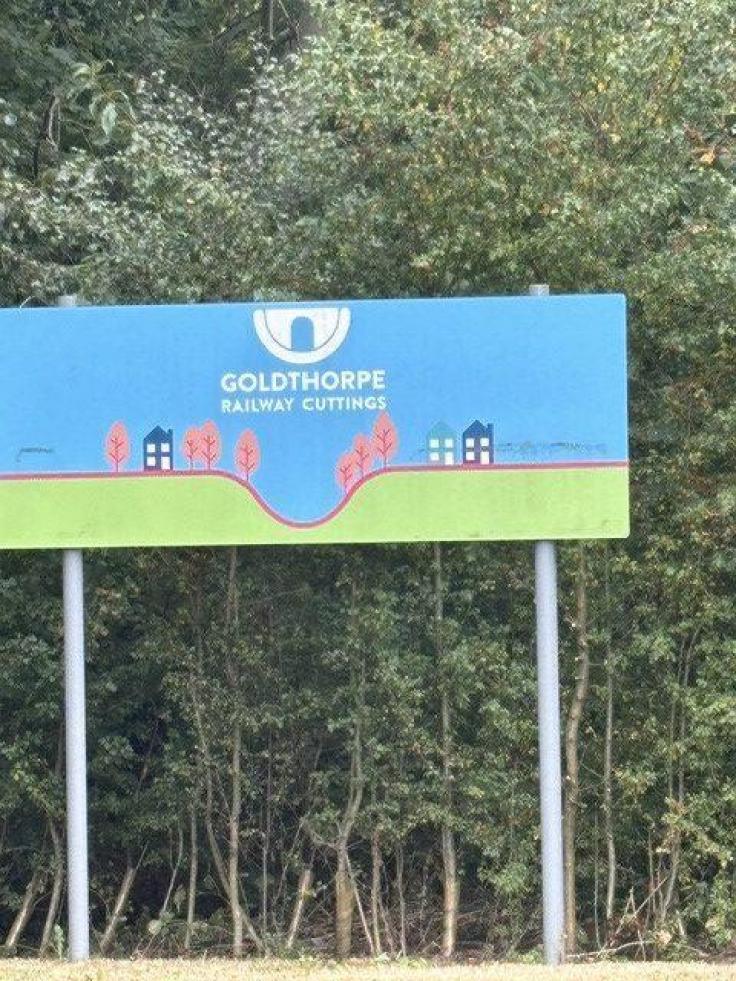 A large rectangular sign on two metal poles reading 'Goldthorpe Railway Cuttings' and green trees behind