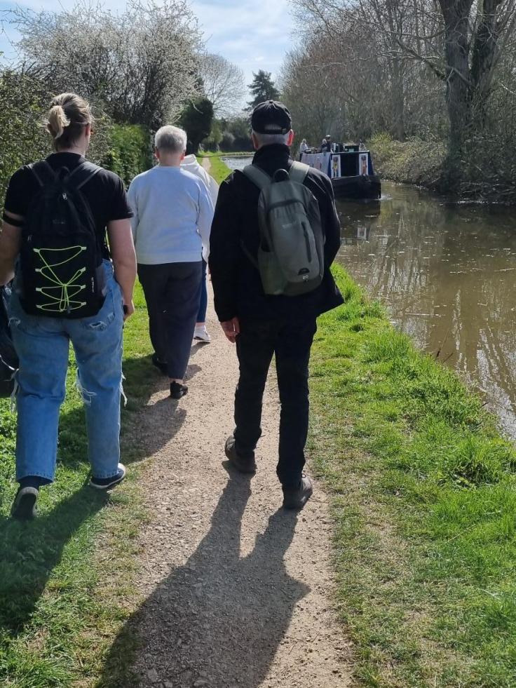People walking along a canal footpath in the sunshine.