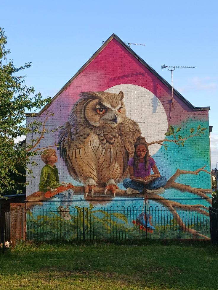 A mural on the gable side of a house featuring a large owl and a small boy sitting on the branch of a tree.