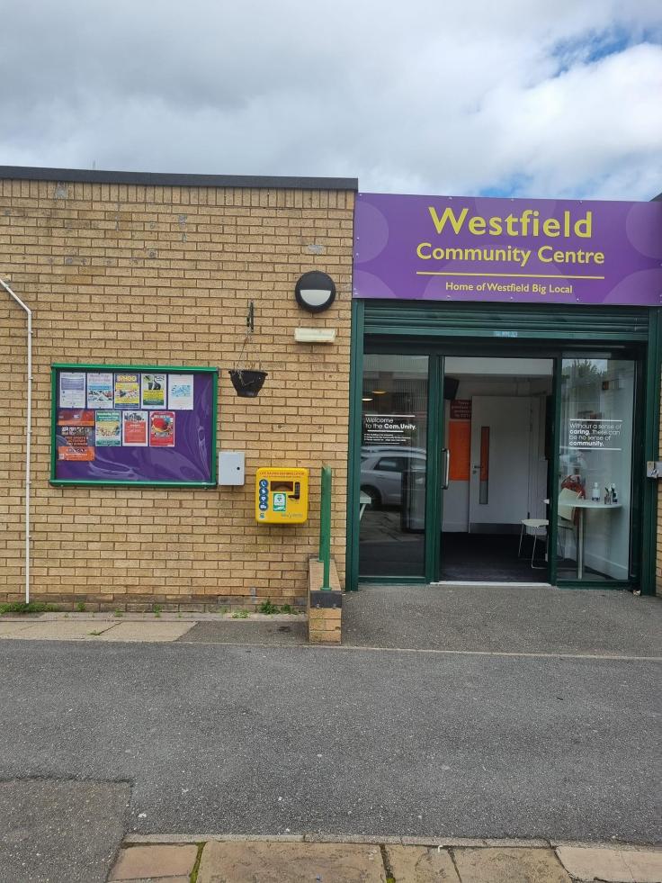 Entrance to the centre, with a noticeboard and defibrillator on the left, and a large purple sign that reads "Westfield Community Centre, Home of Westfield Big Local" above the open entrance doors.