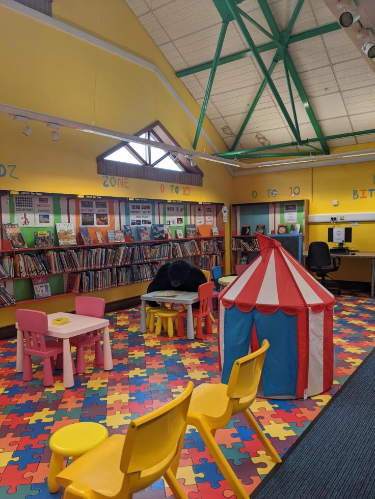 A tall building with green steel rafters, lined with books and featuring a play area with colourful rubber jigsaw flooring.