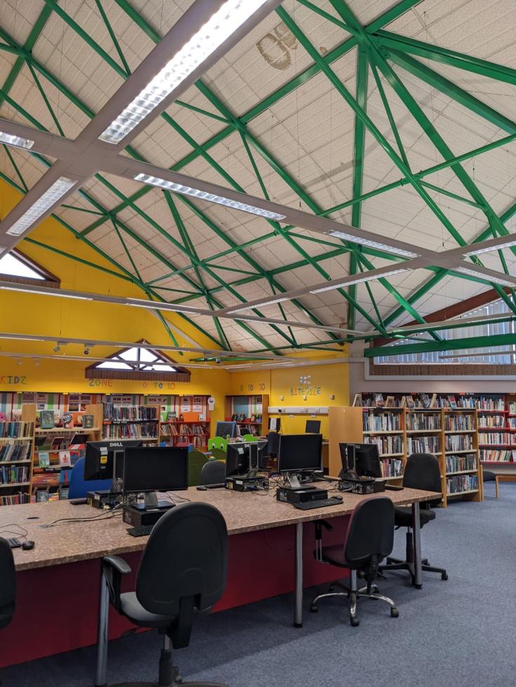 An interior view of a tall building with green rafters, work-spaces with chairs and lined with bookcases.