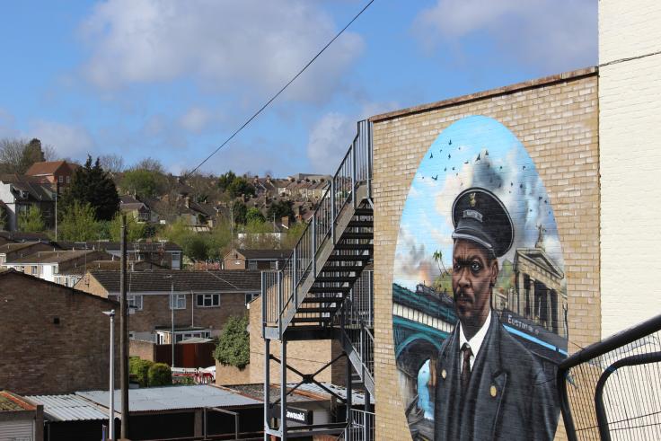 A colourful mural on a brick wall showing a uniformed black railway worker with a mustache, and a steam train traversing a viaduct.
