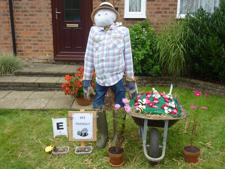 A scarecrow in the front garden of a house, with two button eyes, a straw hat, a plaid shirt and a wheelbarrow of flowers and bees.