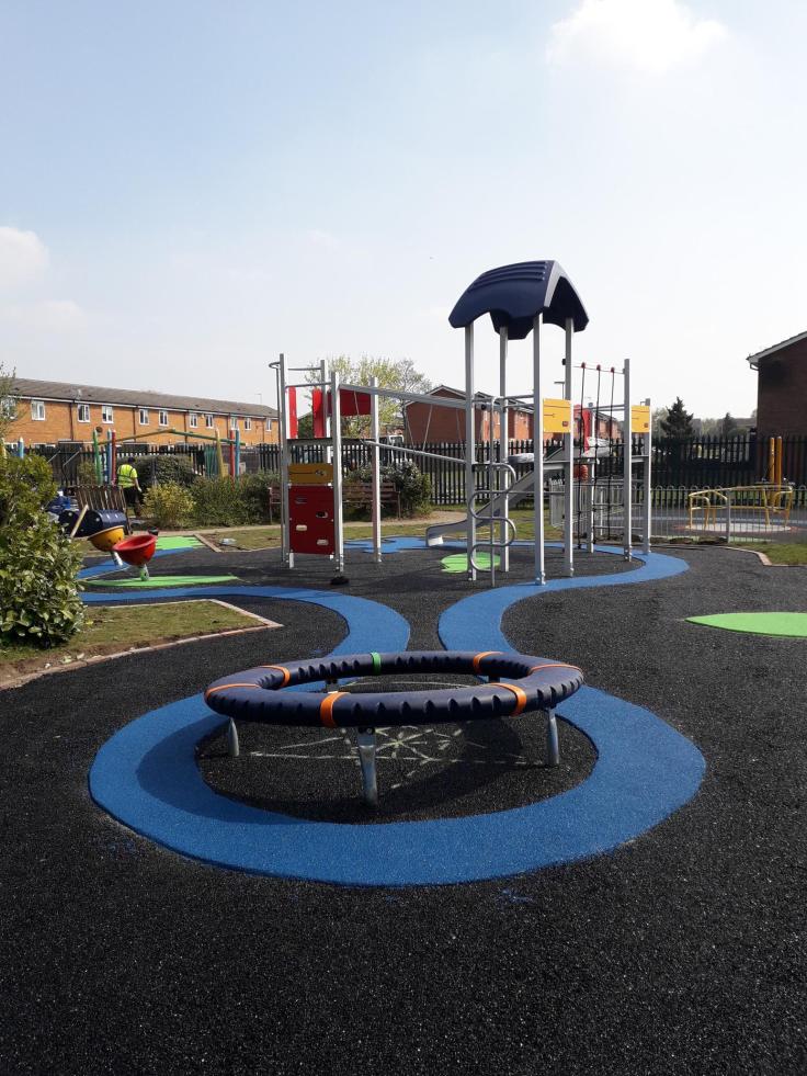 A children's outdoor play park with a climbing frame and other colourful play equipment. There are blue and green patterns painted onto the black asphalt. Rows of terraced houses can be seen behind the park fence.
