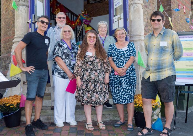 Seven smiling people, on the steps of a building which is decorated for a celebration.