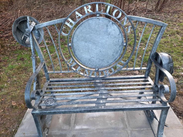 A grey metal bench in a woodland area with a circular design on the back reading 'Charltons memorial'.