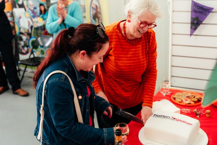 A younger woman in a denim jacket with reddish hair looks down while laughing, holding a glass of white wine. Next to her, an older woman with white hair, red glasses, and a bright orange striped sweater beams with delight. They're standing at a red-covered table that has a white cake decorated with "Fratton Together" text and a plate of biscuits. In the background, there are community banners and purple bunting, and part of a woman in a blue cardigan clapping.