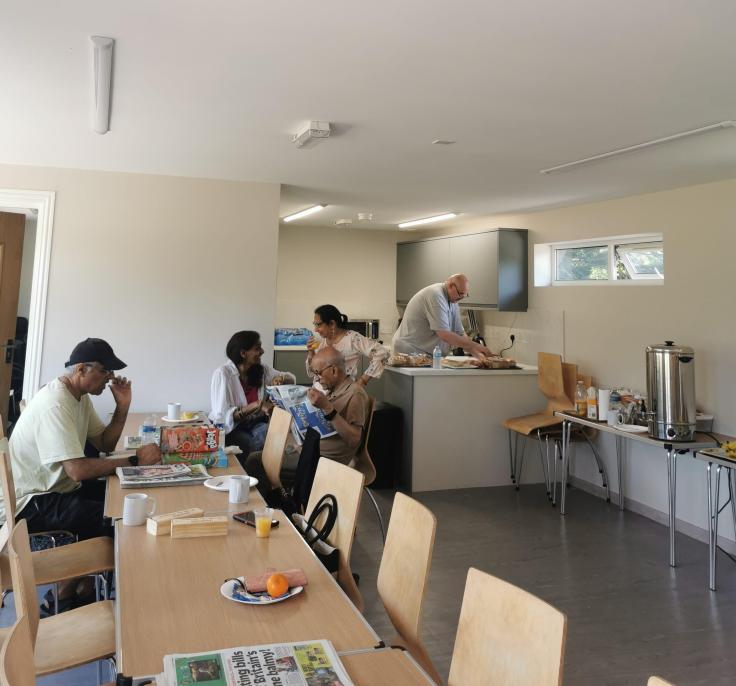 Two women talking, one man reading a newspaper and another man setting up a board game on a long wooden table.