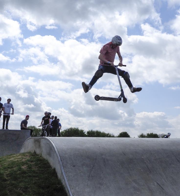 Looking up a concrete skate ramp with a young person mid-air on a scooter, silhouetted against a cloudy sky.