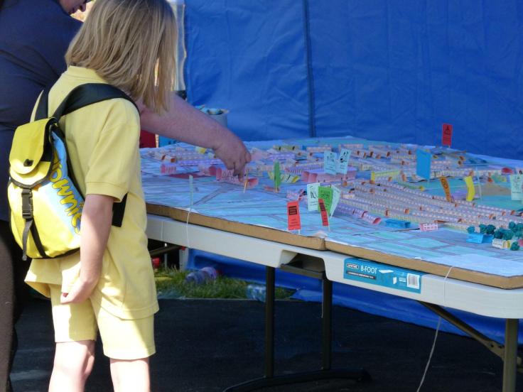 A little girl wearing yellow shorts, a yellow tshirt and a yellow backpack has her back to the camera while looking at a 3D map of housing in the area set on a trestle table. A woman with a blue tshirt and glasses stands next to her, pointing at something on the map. A blue tarpaulin can be seen in the background.
