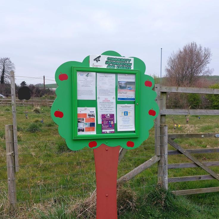 A noticeboard in the shape of a tree by a wooden gate leading into a field.