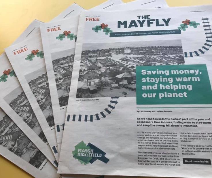 Newspapers titled 'The Mayfly' fanned out on a table.