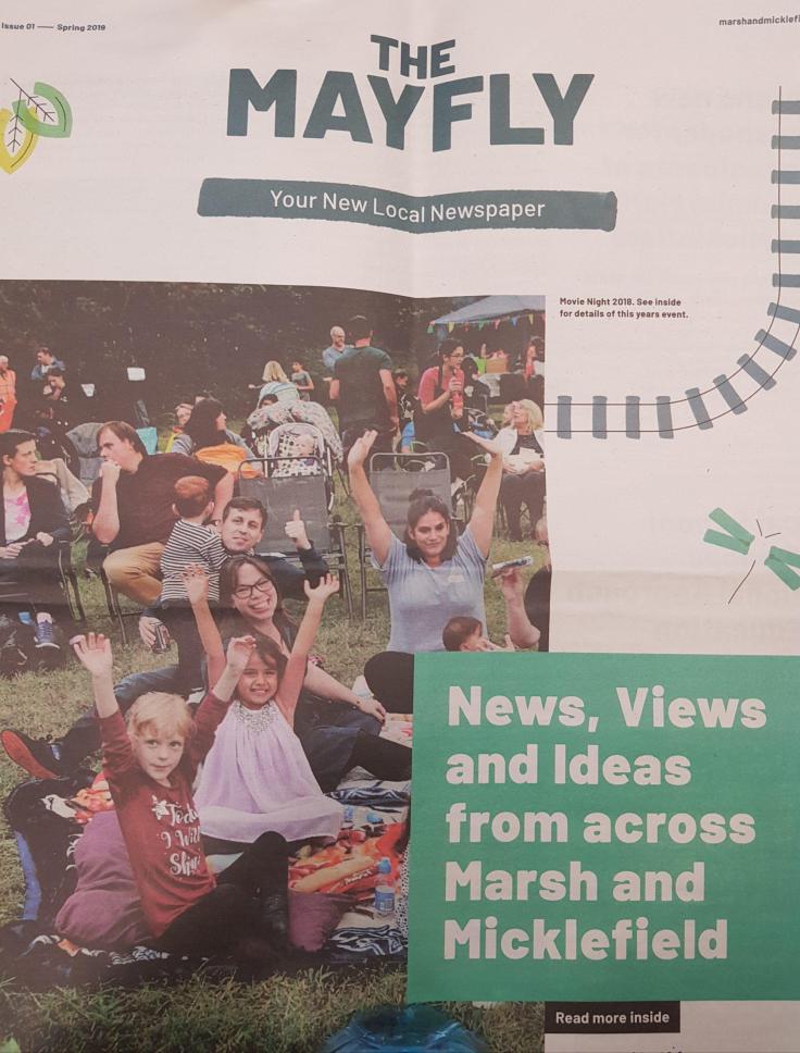 A close-up of The Mayfly local newspaper front page shows a group of smiling children, women and a man with their arms up, smiling. They are sitting on blankets on the grass, with people in camping chairs, a gazebo with bunting, a child in a pram, and mature trees in the background. A green text box reads "News, views and ideas from across Marsh and Micklefield", with the subheading "Read more inside" underneath. There is a geometric navy ladder and a geometric green dragonfly and leaves in the background.