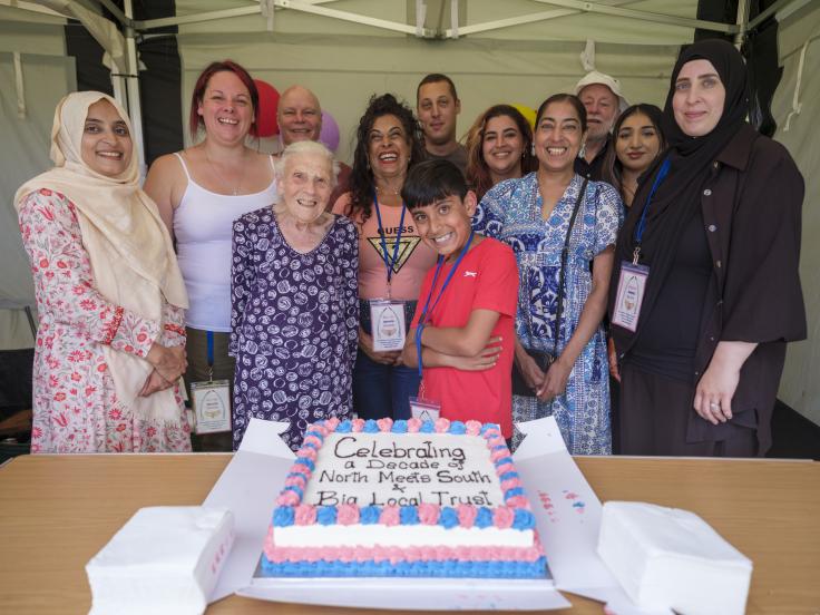 A group of diverse people by ethnicity, age and gender smile at the camera. In front of them is a square sheet cake on a wooden table. The cake has blue and pink icing on the edge of it, and in the middle reads "Celebrating a decade of North Meets South + Big Local Trust".