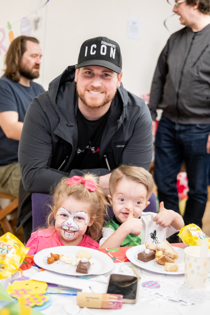 A man wearing a black t-shirt, black jacket and black cap smiles while leaning on the back of a purple chair where two children are sitting and smiling at the camera. The girl on the left wears a pink t-shirt, a pink bow in her hair and her face is painted like a rabbit. The boy on the right has two thumbs up and a green and white football t-shirt. They each have a paper plate in front of them with a selection of sandwiches, cakes and snacks. There are also pencils, a blue and pink paper egg and a chick on the table. Behind them, two more men are in a discussion.