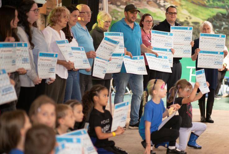 A row of 10 adults of different ages stand holding big cheques, with a row of children kneeling in front of them, also holding big cheques. They are all looking at someone out of frame, probably a photographer.