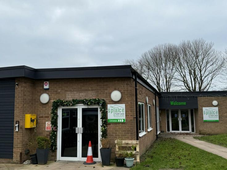 A single-storey brick community centre. It has two sets of white double doors. One has a black sign with green writing on it that reads "Welcome". Two other signs with white backgrounds and green writing say "The Space". There are mature trees without leaves behind the building, and an overcast sky.