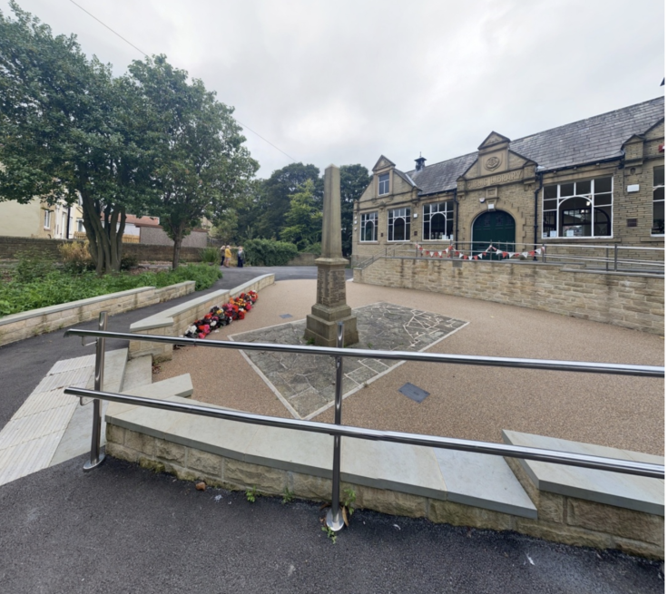 A newly-laid concrete ramp and railing leads to a public square with an obelisk in the middle of it, and a brick-building in the background. There are flowers and a string of red and white bunting in the square. There is also a clump of trees on the left, and overcast sky.