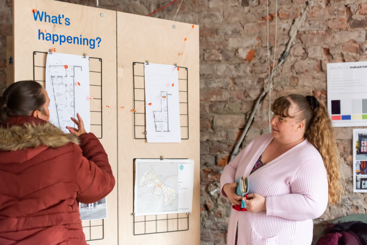 Two people are standing next to a wooden board with plans for the community hub pinned to it. Blue text reads 'What's happening?'. They appear to be discussing what's on the board.
