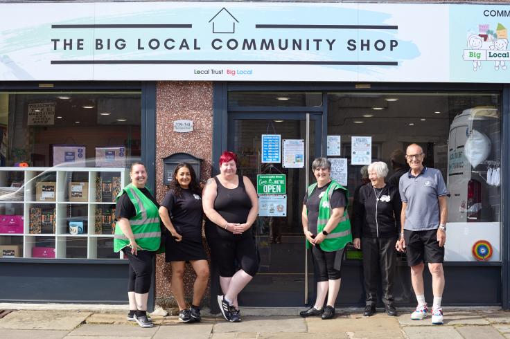 Five women and a man of mixed ages stand outside "The Big Local Community Shop" smiling.