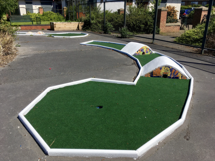 Two mini-golf obstacles with fake grass and white edges are visible against a concrete floor. There are bushes and railings behind and on the right of the image.