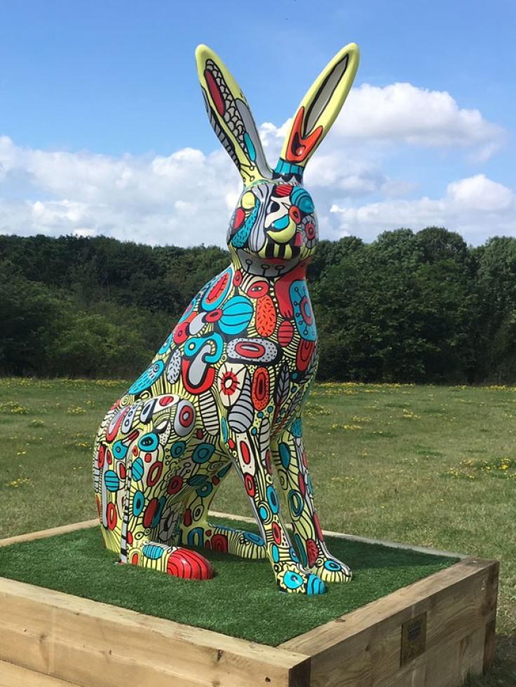 A giant colourful seated rabbit sculpture stands on grass in a wooden plinth. The sculpture is covered in yellow, blue, grey and red shapes and abstract patterns. Behind it is a green field with yellow flowers, mature green trees on the horizon and a blue sky with white clouds above.