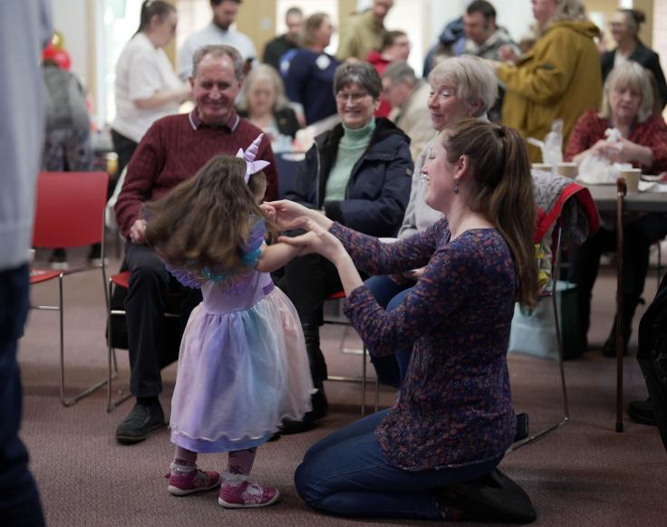 A woman wearing a purple top and jeans sits on the floor while holding the hands of a little girl dancing  in a purple unicorn costume on a red carpet. Three other adults sit on chairs watching them. Blurred in the background are more people standing and talking.