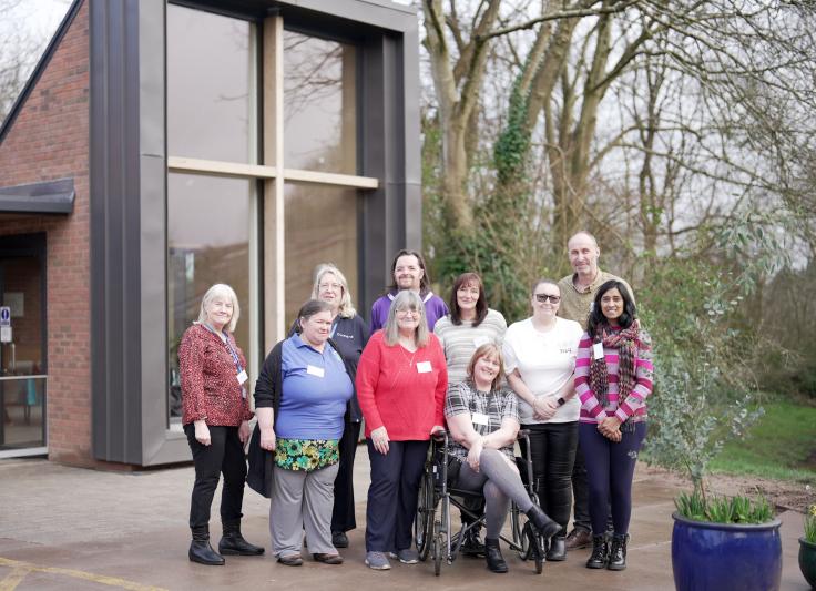 A group of people smiling at the camera, in front of a modern-looking building extension with floor-to-ceiling windows. One young woman is in a wheelchair, the rest are standing. On the right there are trees without leaves.