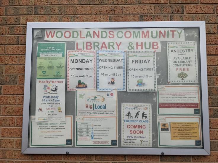 A noticeboard covered in colourful posters with a sign reading 'Woodlands Community Library and Hub'.