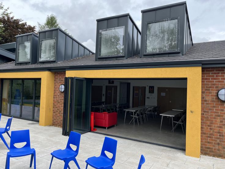 One-storey building with raised sloping skylights and open glass doors showing tables and chairs inside along with blue chairs in a circle outside the building.